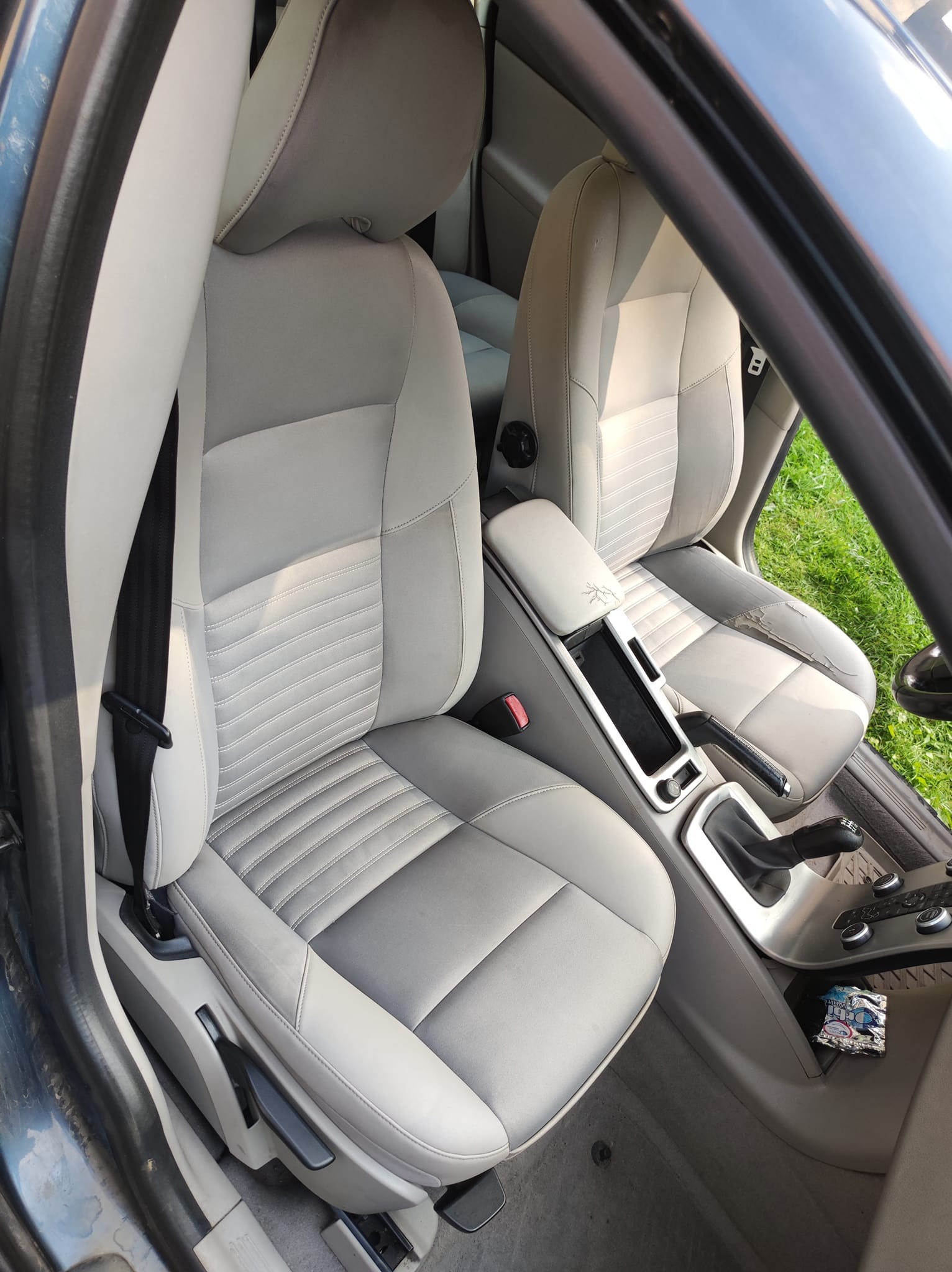 Jasnoszare fotele samochodowe z wyraźnymi śladami zużycia, szczególnie na fotelu pasażera, widoczne przez otwarte drzwi auta na tle zielonej trawy.