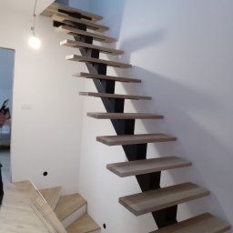 STYL -SPAW - Nowoczesne schody wspornikowe z drewnianymi stopniami i czarną, metalową konstrukcją nośną, zamontowane przy białej ścianie. Widoczny fragment podłogi i niedokończone wykończenie.