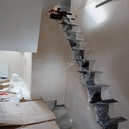 STYL -SPAW - Czarne, metalowe schody wspornikowe, zabezpieczone folią, w trakcie montażu w surowym wnętrzu. Widoczne przygotowania do dalszych prac wykończeniowych.