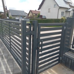 STYL -SPAW - Nowoczesna, pozioma brama wjazdowa z metalowymi profilami w kolorze grafitowym, z widocznymi kluczami w zamku, na tle domów jednorodzinnych i kostki brukowej.