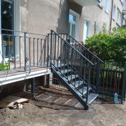 STYL -SPAW - Zewnętrzne schody metalowe z balustradą prowadzące do budynku. Schody z kraty pomostowej, ciemnoszara konstrukcja. Widoczne elementy wykończeniowe i otoczenie.