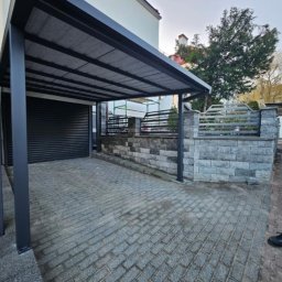 STYL -SPAW - Zadaszenie nad podjazdem z szarej kostki brukowej, wsparte na metalowych słupach. W tle budynek z szarą bramą garażową i ogrodzenie z metalowymi przęsłami.