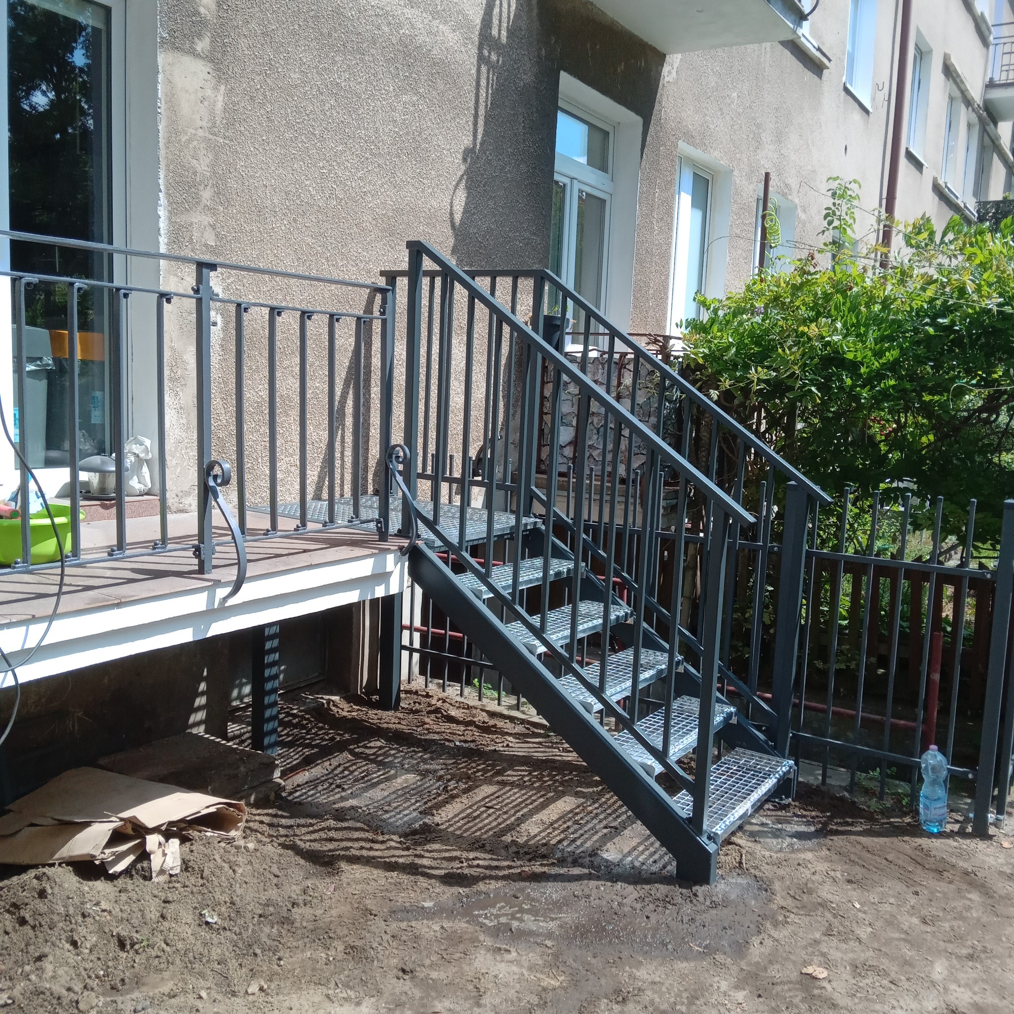 Zewnętrzne schody metalowe z balustradą prowadzące do budynku. Schody z kraty pomostowej, ciemnoszara konstrukcja. Widoczne elementy wykończeniowe i otoczenie.