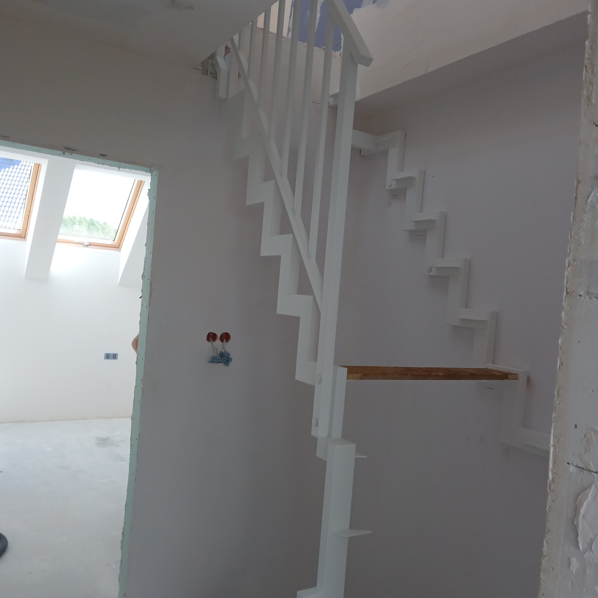 Białe schody z balustradą w surowym wnętrzu, widoczne okno dachowe i instalacja elektryczna. Minimalistyczny design schodów.