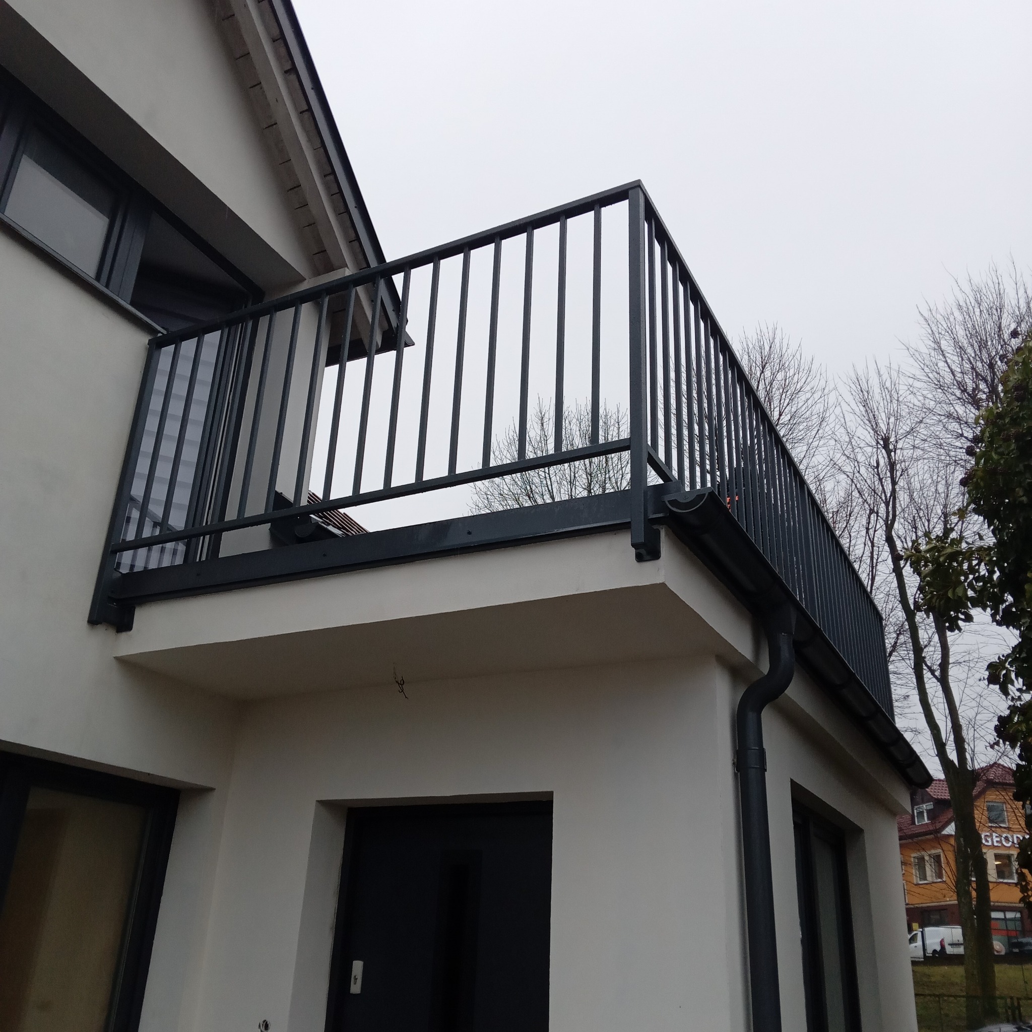 Nowoczesna, grafitowa balustrada balkonowa z pionowymi szczeblami, zamontowana na białym budynku. Widok z dołu, podkreślający minimalistyczny design i solidne wykonanie.