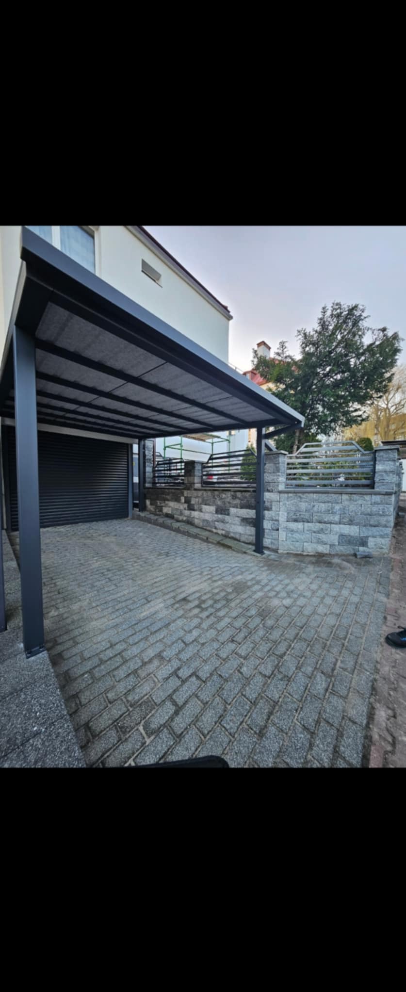 Zadaszenie nad podjazdem z szarej kostki brukowej, wsparte na metalowych słupach. W tle budynek z szarą bramą garażową i ogrodzenie z metalowymi przęsłami.