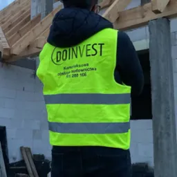 Osoba w kasku i kamizelce odblaskowej z logo 'DOINVEST' na tle budowy domu w stanie surowym otwartym, widoczne drewniane elementy konstrukcji dachu.
