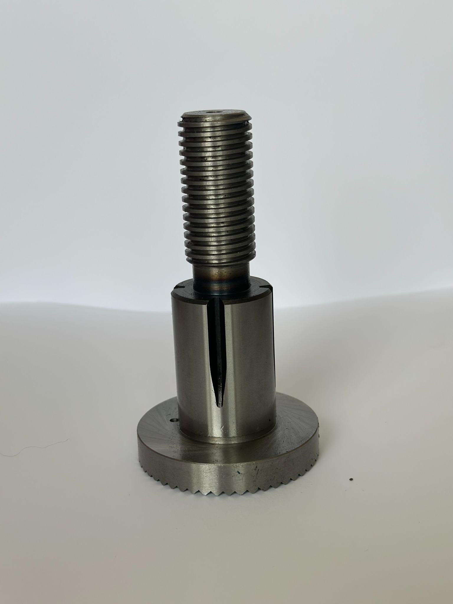 Metalowy uchwyt tokarski z gwintowanym trzpieniem i promieniowym nacięciem na cylindrycznej części, sfotografowany na jasnym tle.