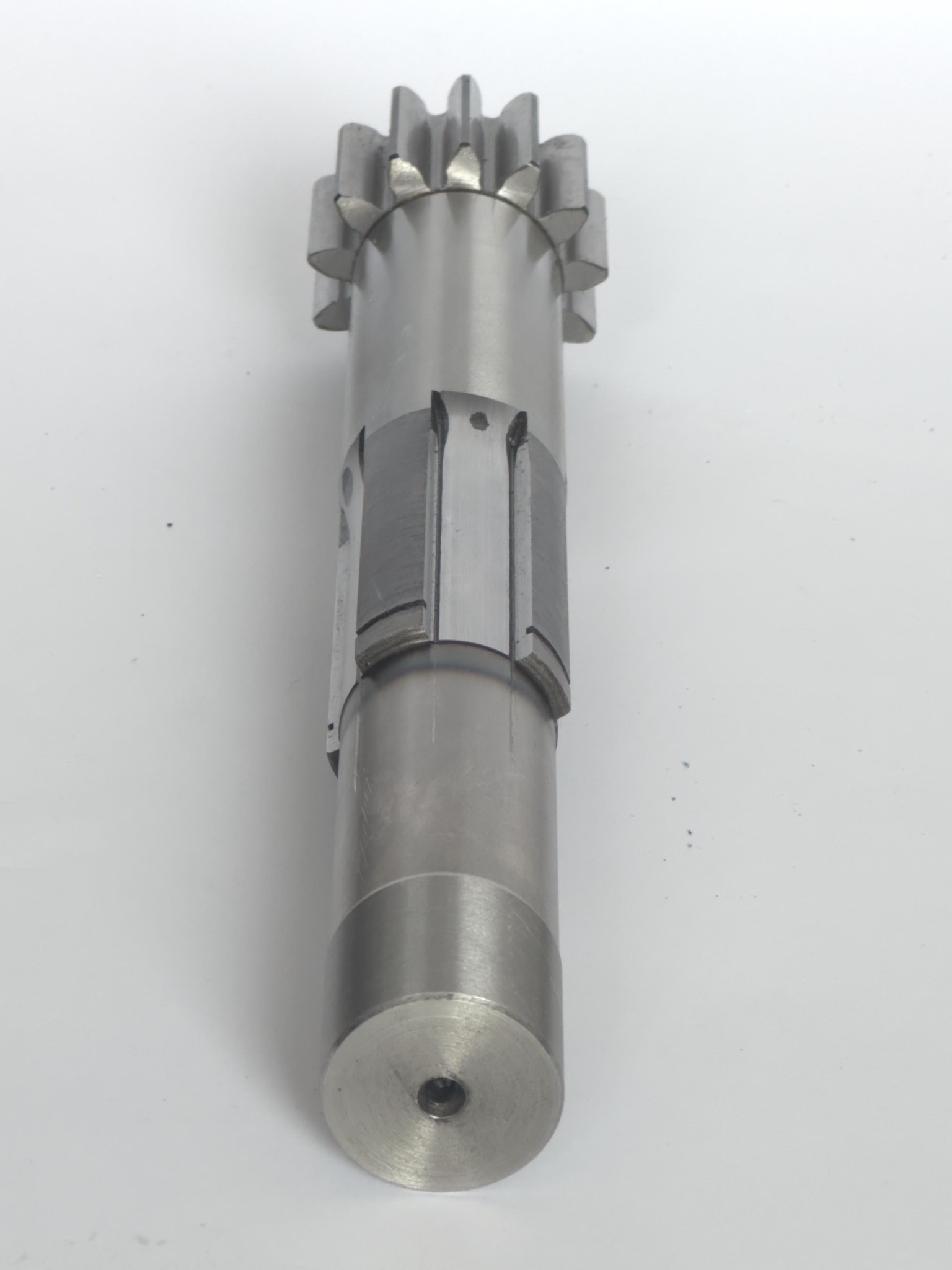 Metalowy wał z zębatką i frezowanymi rowkami, widoczne ślady obróbki, na jednolitym jasnym tle.