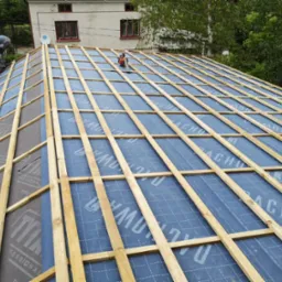 Konstrukcja dachu z drewnianych łat i kontrłat, pokryta niebieską membraną dachową z nadrukowanym napisem, pracownik na krawędzi dachu w tle.