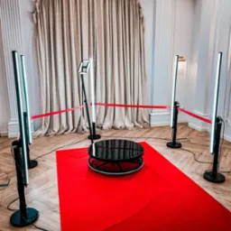 Obracana platforma w otoczeniu lamp LED i czerwonego dywanu, całość odgrodzona czerwoną taśmą, ustawiona w eleganckim wnętrzu z kolumnami i drapowanymi zasłonami.