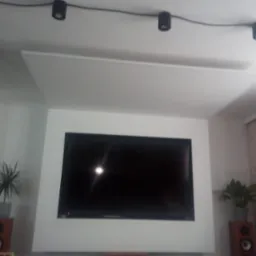 Nowoczesny salon z podwieszanym sufitem, oświetleniem punktowym i telewizorem w obudowie, rośliny doniczkowe na głośnikach.