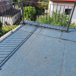 Niebieski dach płaski pokryty papą na balkonie w Gdańsku, z metalową balustradą i widokiem na zieleń w tle.