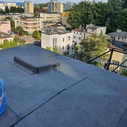 Dachy Czajkowscy - Michał Czajkowski - Widok z góry na płaski dach pokryty papą, z niebieską butlą gazową w lewym dolnym rogu i zabudową wentylacyjną pośrodku, w tle zabudowa miejska i zieleń