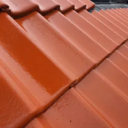 Błyszcząca, nowa dachówka ceramiczna w odcieniach czerwieni, ułożona równo na skośnym dachu, z widocznymi śladami wilgoci lub świeżo nałożonej impregnacji.