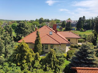 Dom jednorodzinny z nowym, pomarańczowym dachem z dachówki ceramicznej, widoczny z góry, otoczony zielenią drzew i krzewów, przy słonecznej pogodzie.