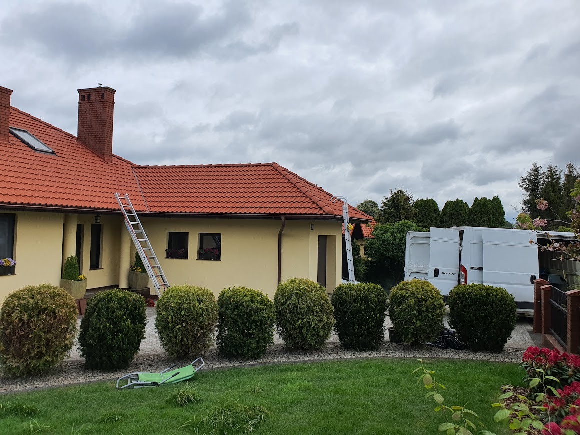 Dom jednorodzinny z czerwoną dachówką, drabiny opierają się o dach, biały samochód dostawczy zaparkowany obok, na pierwszym planie rząd kulistych krzewów i zielony leżak.