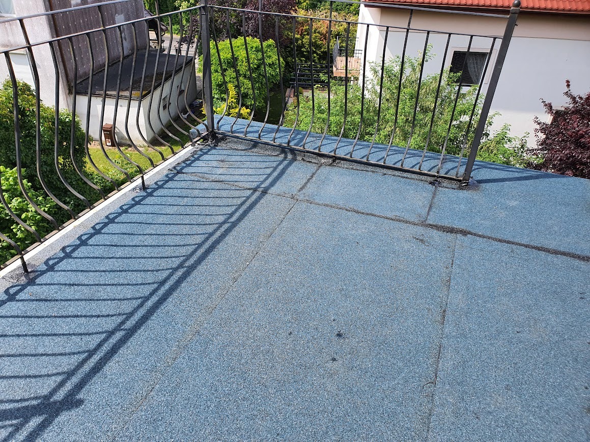 Niebieski dach płaski pokryty papą na balkonie w Gdańsku, z metalową balustradą i widokiem na zieleń w tle.