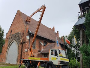 Pomarańczowy podnośnik koszowy na tle ceglanego kościoła z widocznymi ubytkami w murze i polską flagą powiewającą w tle. Ramię podnośnika sięga dachu budynku.