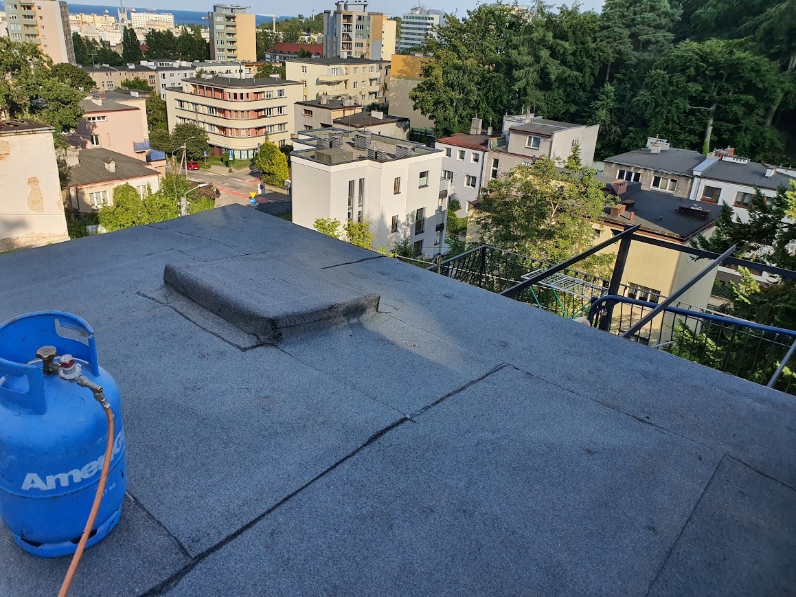 Widok z góry na płaski dach pokryty papą, z niebieską butlą gazową w lewym dolnym rogu i zabudową wentylacyjną pośrodku, w tle zabudowa miejska i zieleń