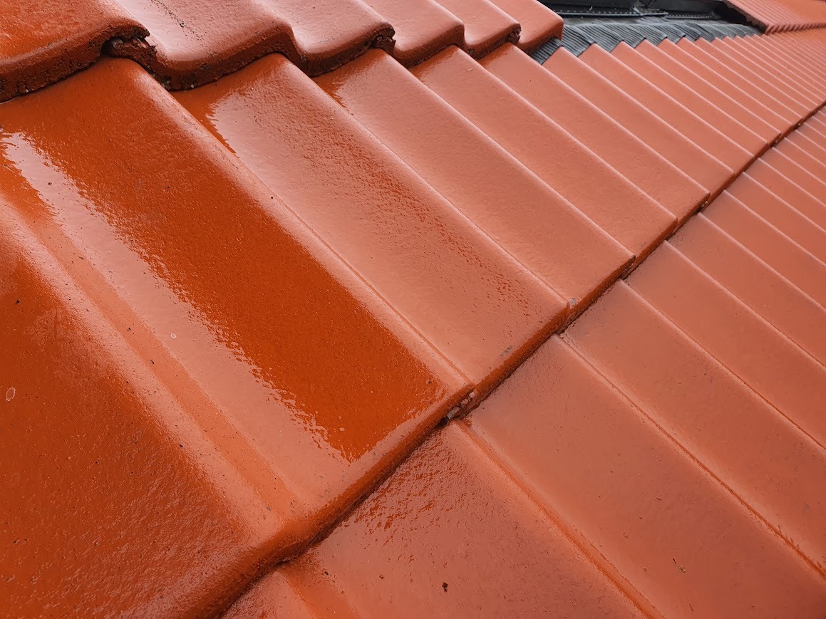 Błyszcząca, nowa dachówka ceramiczna w odcieniach czerwieni, ułożona równo na skośnym dachu, z widocznymi śladami wilgoci lub świeżo nałożonej impregnacji.