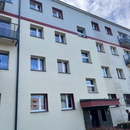 Profada Filip Adamiec - Wielopiętrowy budynek mieszkalny z kremową i szarą elewacją, widoczne okna z czerwonymi obramowaniami, balkony i wejście do budynku otoczone krzewami.