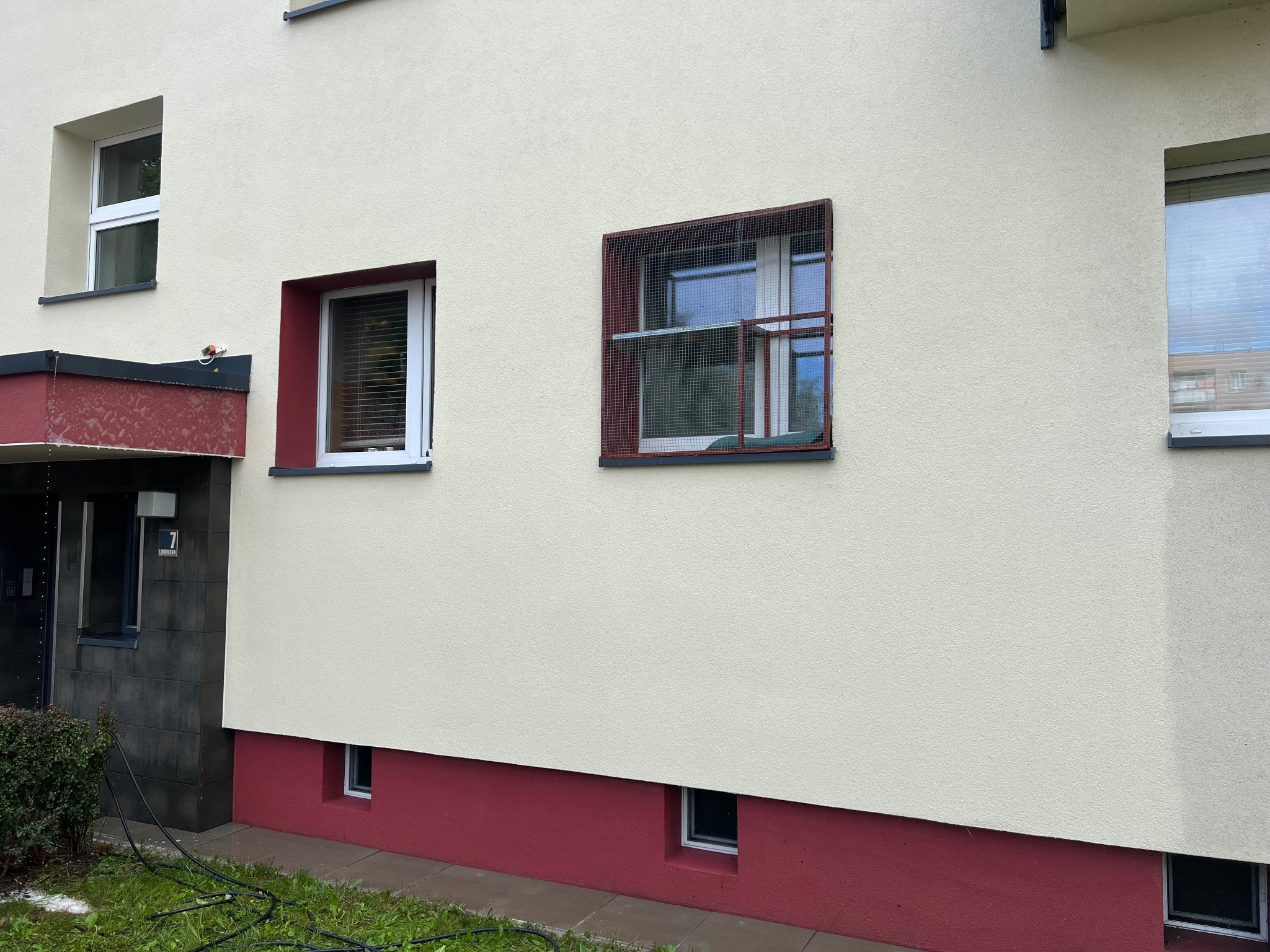 Elewacja budynku wielorodzinnego z oknami, jedno z nich zabezpieczone metalową siatką ochronną, dolna część pomalowana na bordowo, widoczne przewody na trawniku.