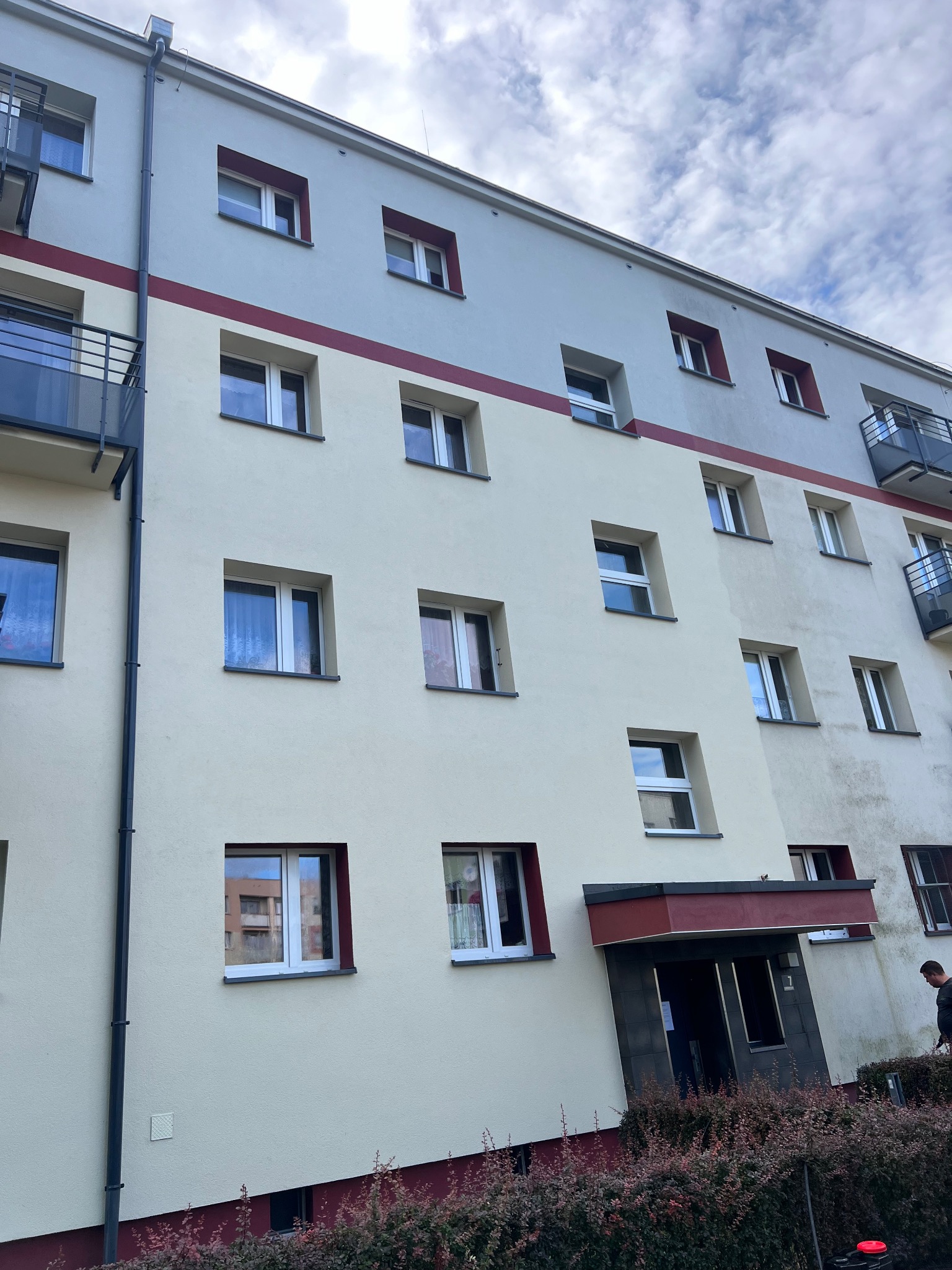 Wielopiętrowy budynek mieszkalny z kremową i szarą elewacją, widoczne okna z czerwonymi obramowaniami, balkony i wejście do budynku otoczone krzewami.