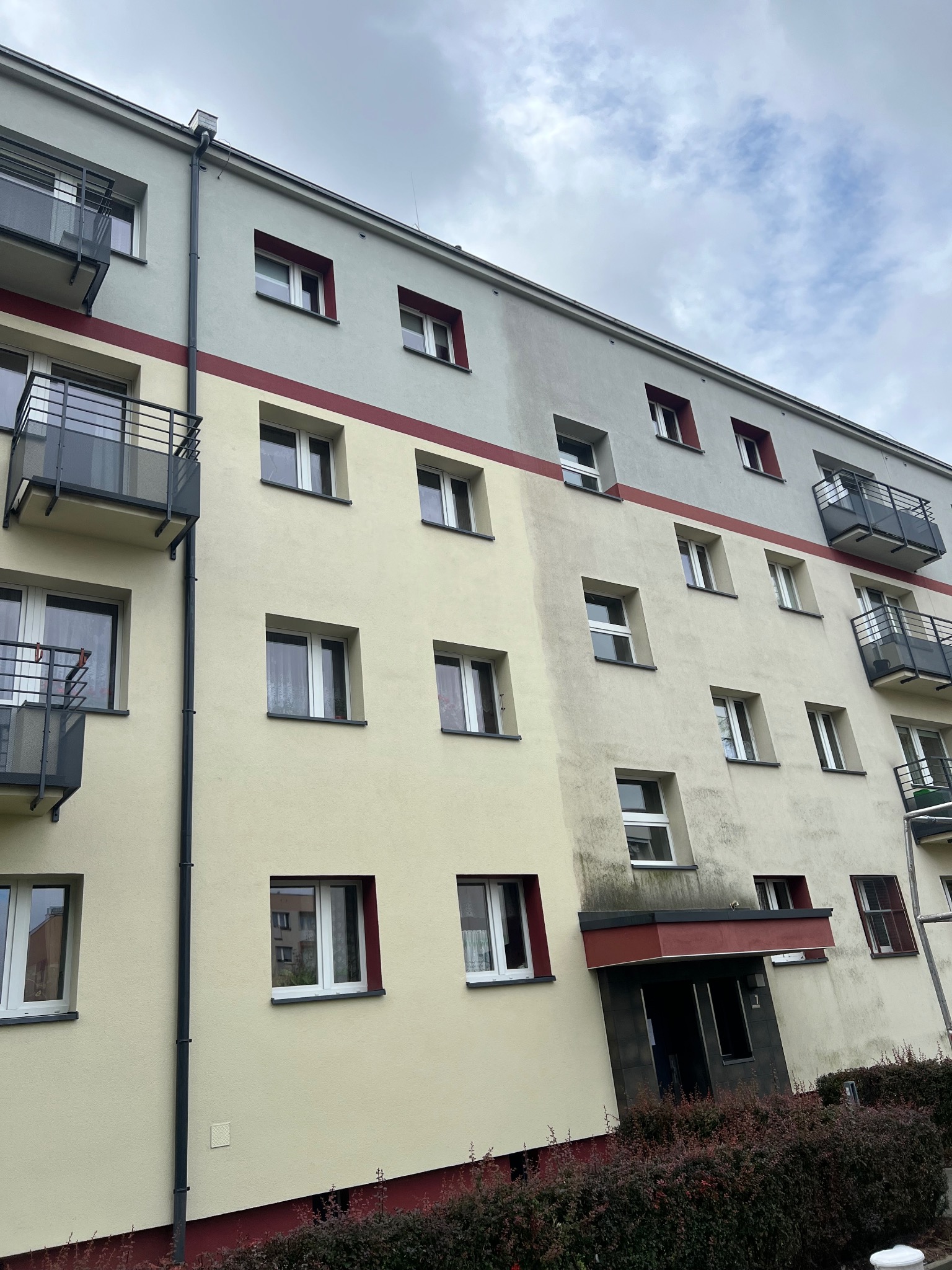 Wielopiętrowy budynek mieszkalny z balkonami, z widocznymi śladami zabrudzeń na elewacji, zwłaszcza nad wejściem i pod oknami.