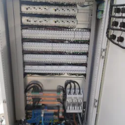 Otwarta szafa elektryczna z widocznymi bezpiecznikami, przewodami i elementami łączeniowymi, uporządkowane okablowanie w kolorze niebieskim i czarnym, szyny miedziane.