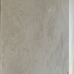 NOOK WYKOŃCZENIA WNĘTRZ KRYSTIAN ZYCH - Ściana pokryta szarą, dekoracyjną strukturą. Widoczne nieregularne wzory i faktura. Ujęcie z boku, ukazujące grubość nałożonego materiału. Drobne nierówności i cieniowania.