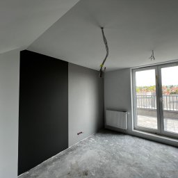 NOOK WYKOŃCZENIA WNĘTRZ KRYSTIAN ZYCH - Wnętrze pokoju w trakcie malowania: fragment ściany pomalowany na czarno, reszta na szaro, surowa podłoga, wystające kable z sufitu, grzejnik pod oknem balkonowym z widokiem na dachy budynków.