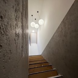 NOOK WYKOŃCZENIA WNĘTRZ KRYSTIAN ZYCH - Klatka schodowa w trakcie prac wykończeniowych; widoczne surowe schody, ściany z efektem dekoracyjnym i nowoczesne, kuliste lampy sufitowe.