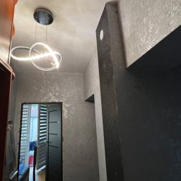 NOOK WYKOŃCZENIA WNĘTRZ KRYSTIAN ZYCH - Wnętrze korytarza z dekoracyjną, srebrzystą fakturą na ścianach, nowoczesną lampą sufitową w kształcie pierścieni oraz czarną ościeżnicą drzwiową.