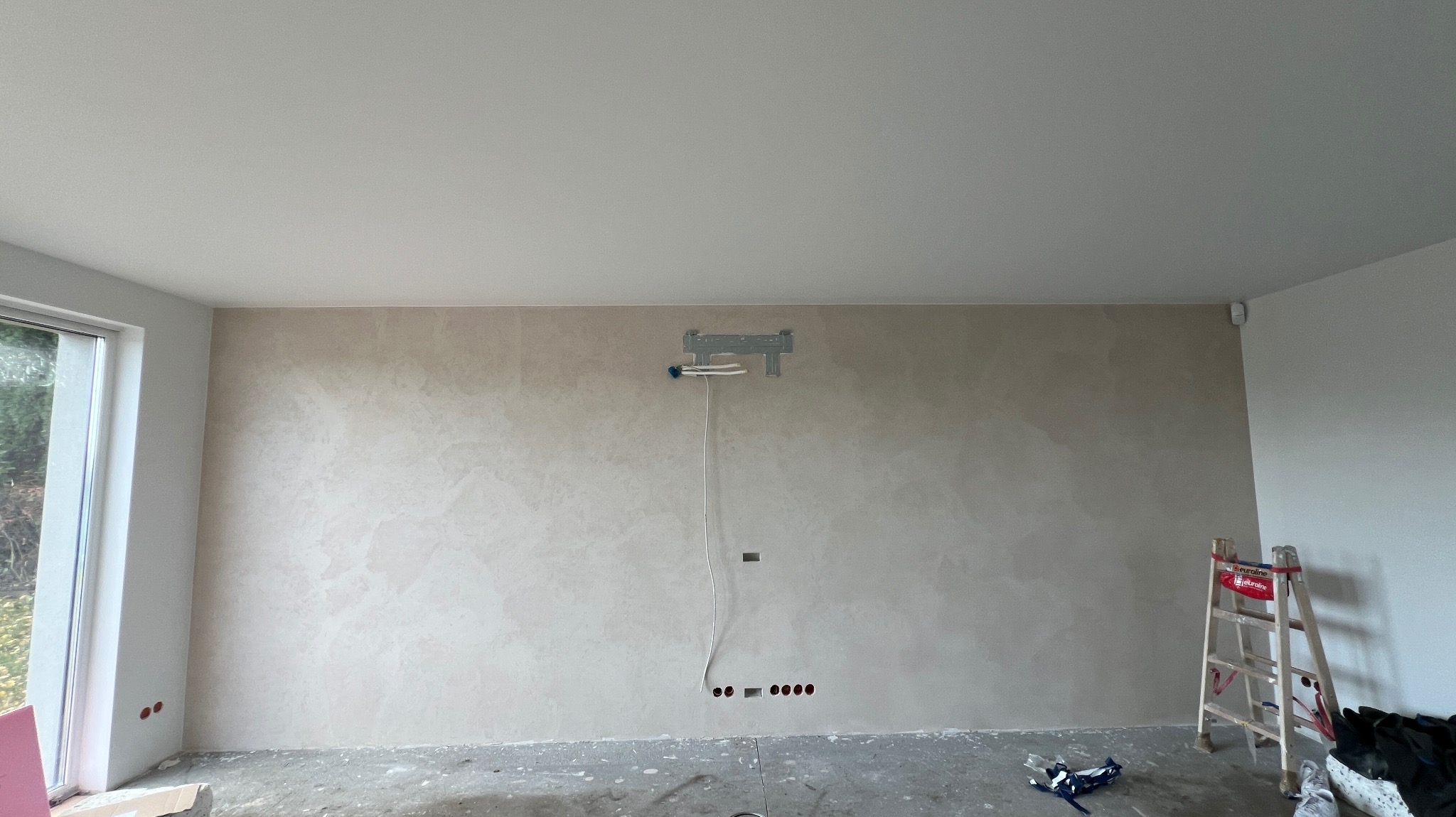 Pokój po szpachlowaniu z fakturą na ścianie, widoczne przygotowania pod montaż TV, drabina oparta o ścianę. Podłoga zabezpieczona folią malarską.