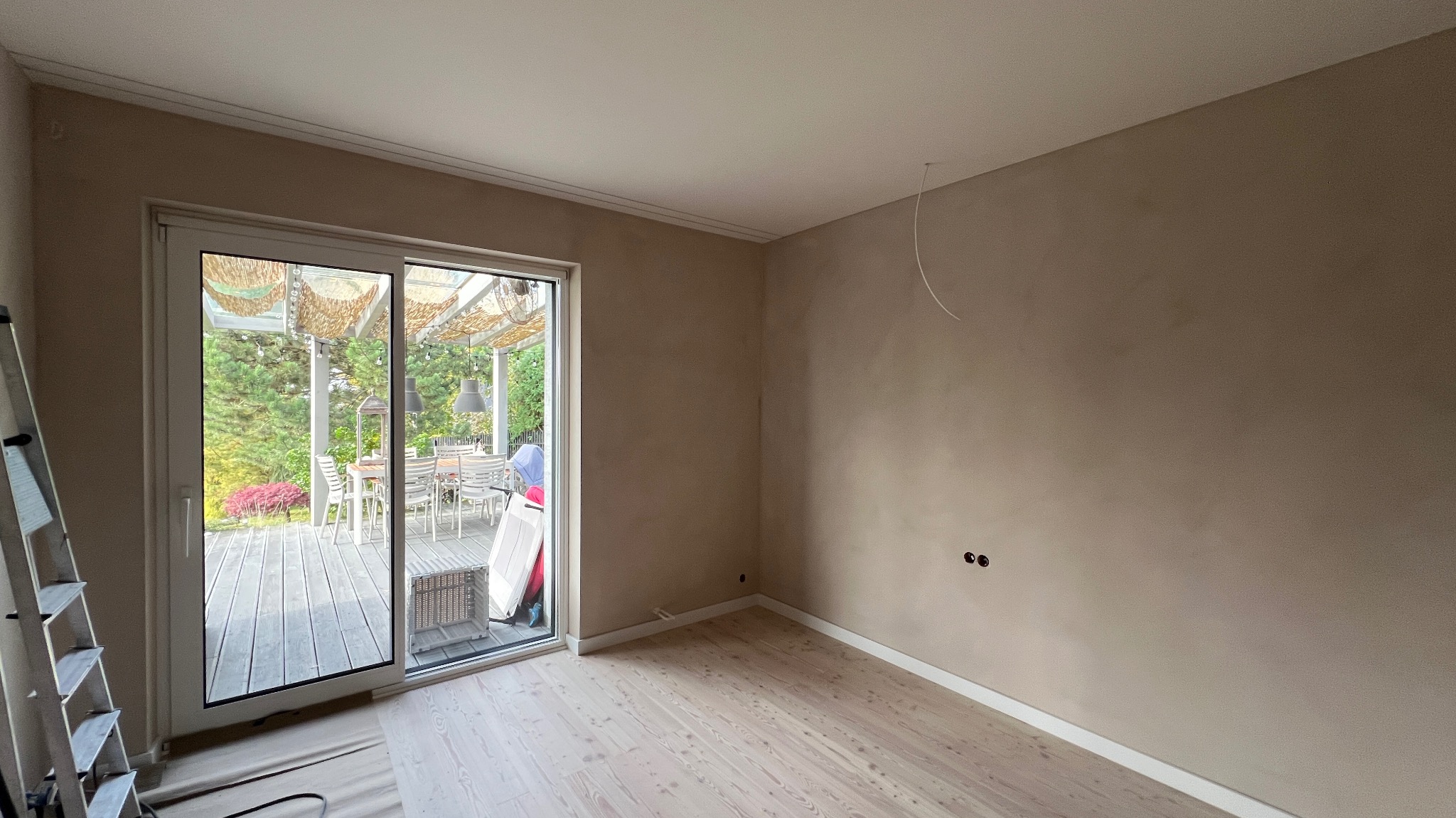 Pokój po szpachlowaniu i malowaniu na beżowo, z widocznymi gniazdkami elektrycznymi, jasną podłogą i drabiną opartą o przesuwne drzwi balkonowe.