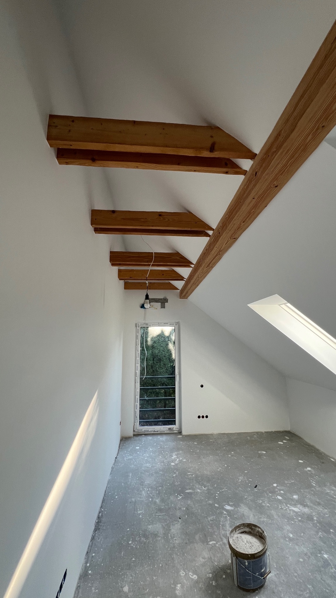 Poddasze po remoncie: białe ściany, drewniane belki stropowe, okno dachowe i zwykłe. Na podłodze resztki farby, stoi wiadro. Widoczne punkty elektryczne.
