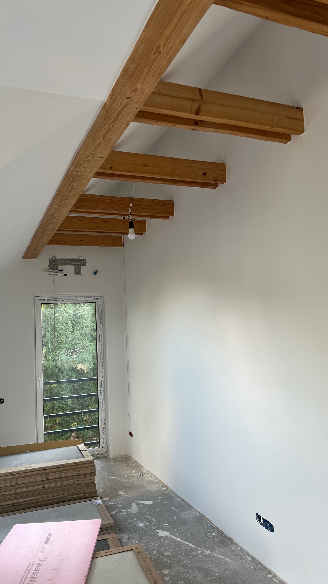 Jasne wnętrze poddasza z drewnianymi belkami stropowymi, świeżo pomalowanymi ścianami i oknem z widokiem na zieleń. Na podłodze widoczne płyty izolacyjne.