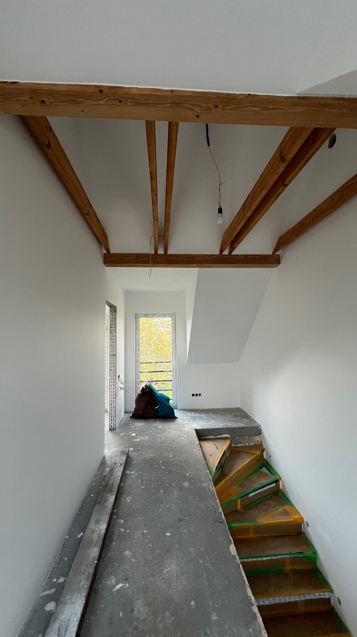 Wnętrze w trakcie wykończenia: białe ściany, drewniane belki stropowe, schody zabezpieczone folią, worki z materiałami budowlanymi. Widoczne przygotowania do dalszych prac.