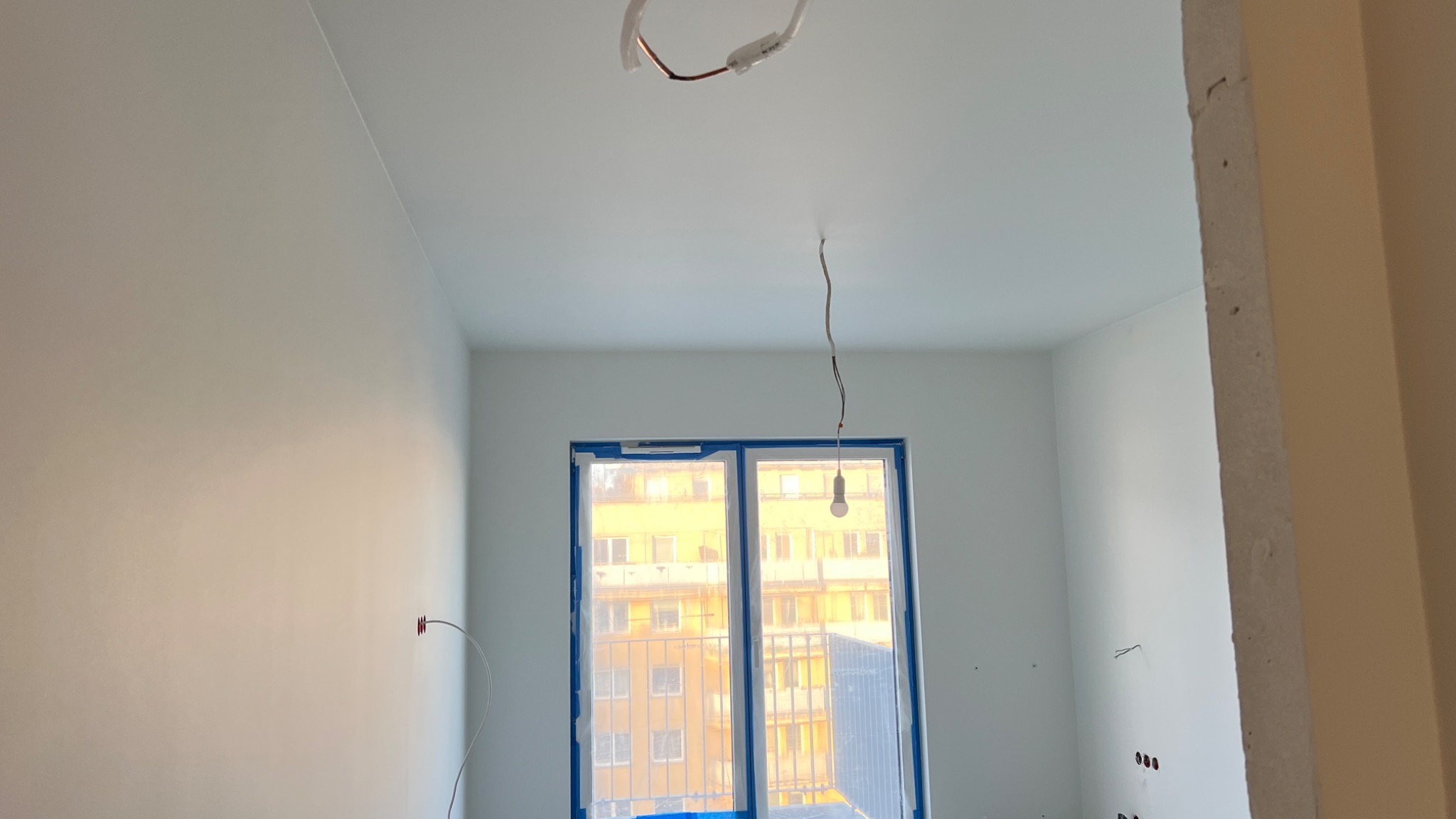 Wnętrze po malowaniu na biało, z widocznymi przewodami elektrycznymi i oknem zabezpieczonym folią malarską. Ściany gładkie, sufit pomalowany. Widoczne gniazdka elektryczne.