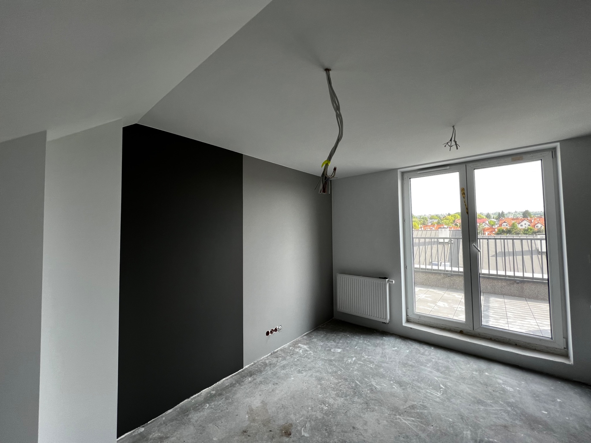 Wnętrze pokoju w trakcie malowania: fragment ściany pomalowany na czarno, reszta na szaro, surowa podłoga, wystające kable z sufitu, grzejnik pod oknem balkonowym z widokiem na dachy budynków.