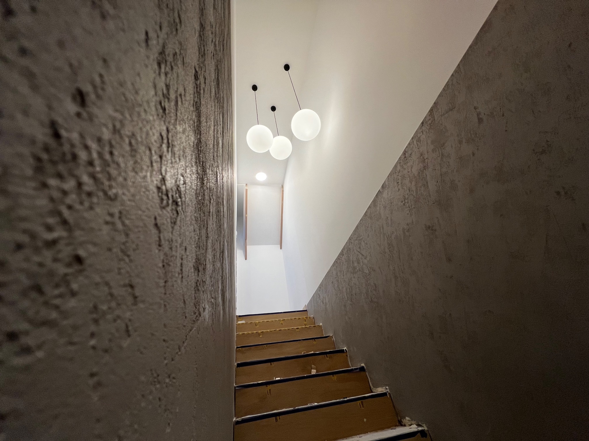 Klatka schodowa w trakcie prac wykończeniowych; widoczne surowe schody, ściany z efektem dekoracyjnym i nowoczesne, kuliste lampy sufitowe.