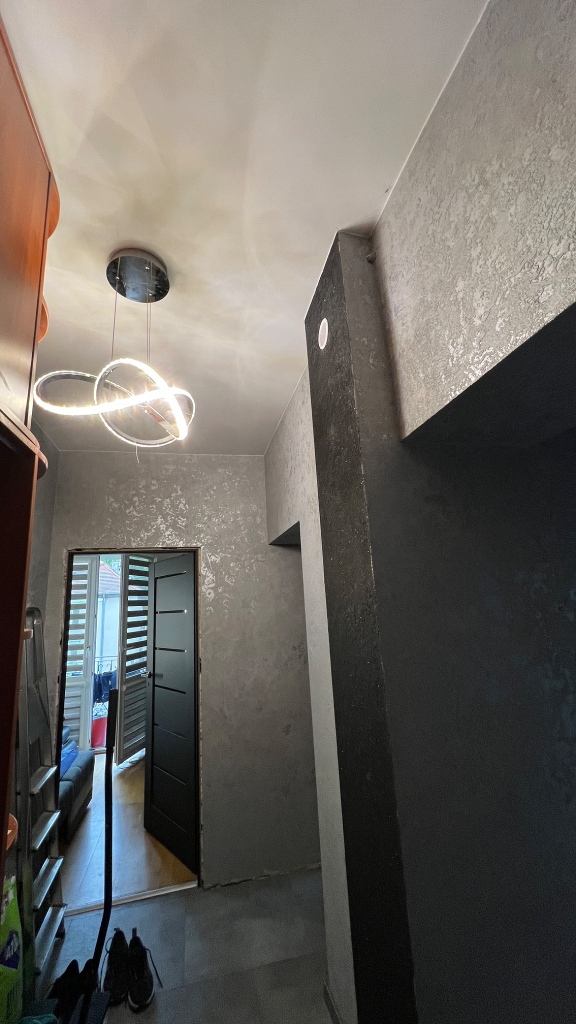 Wnętrze korytarza z dekoracyjną, srebrzystą fakturą na ścianach, nowoczesną lampą sufitową w kształcie pierścieni oraz czarną ościeżnicą drzwiową.