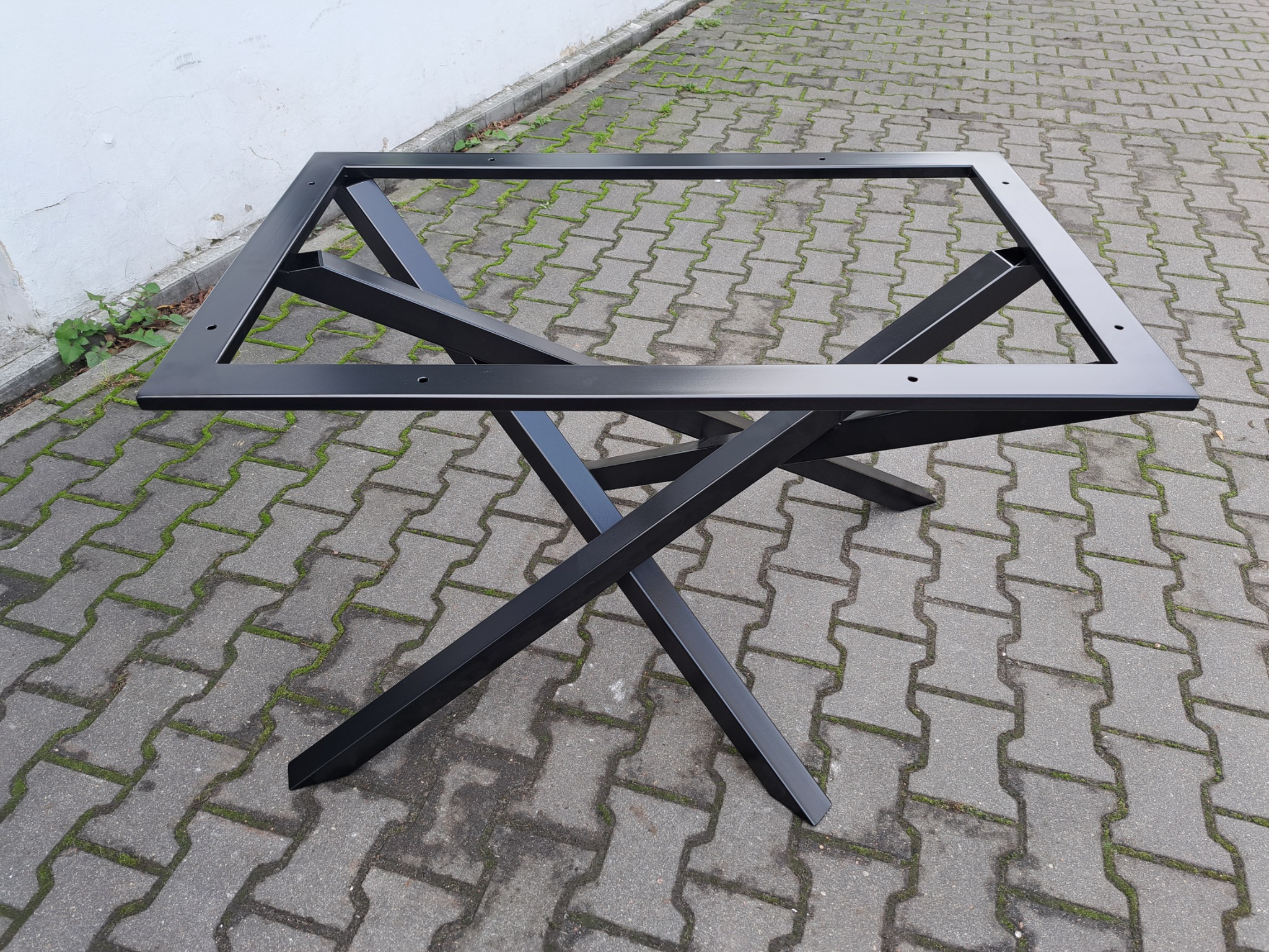 Czarna, metalowa podstawa stołu z krzyżowymi nogami i ramą mocującą blat, sfotografowana na brukowanej nawierzchni w Warszawie.