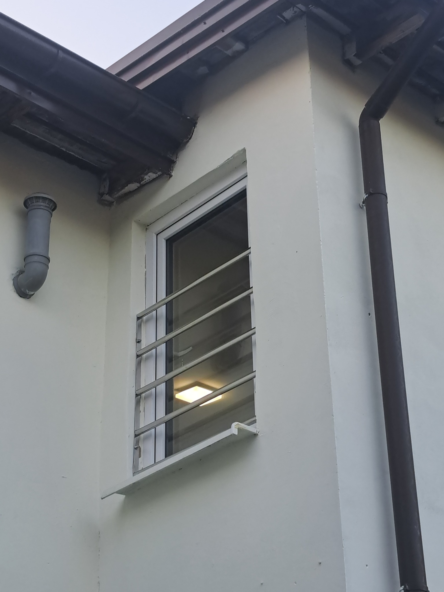 Okratowane okno na zewnątrz budynku z widoczną rurą spustową i wentylacyjną. Stalowe pręty zabezpieczają okno.