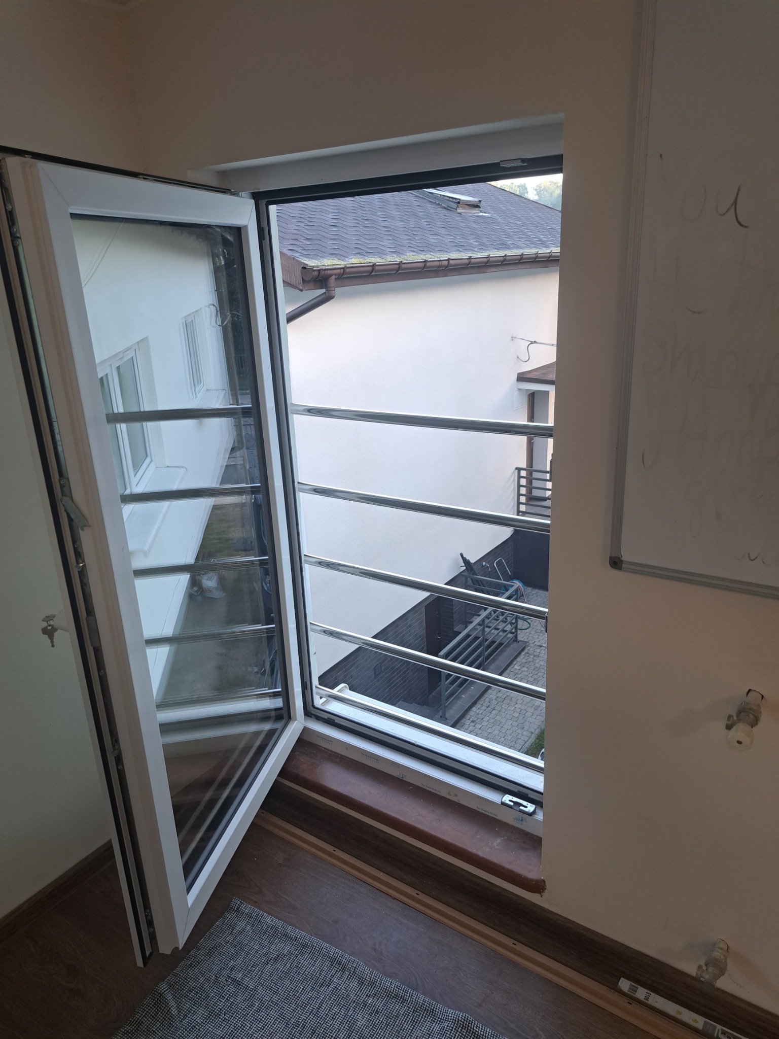 Otwarta okiennica z poziomymi, metalowymi prętami zabezpieczającymi, widok na dziedziniec z metalową balustradą i budynki w tle.