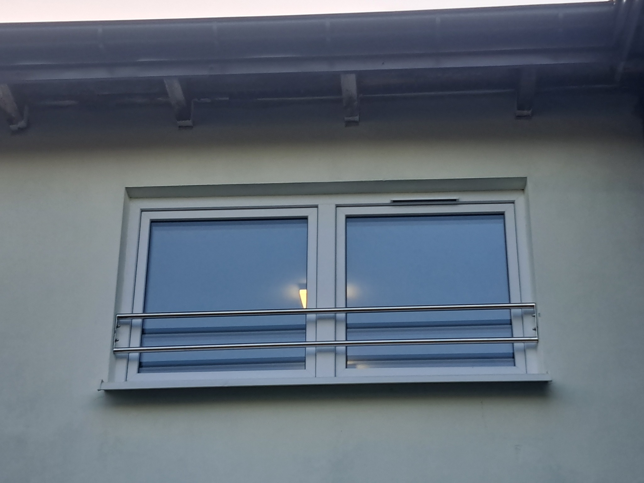 Dwa poziome, nierdzewne zabezpieczenia okienne zamontowane na oknie z białym profilem, widoczne na tle jasnej elewacji budynku.