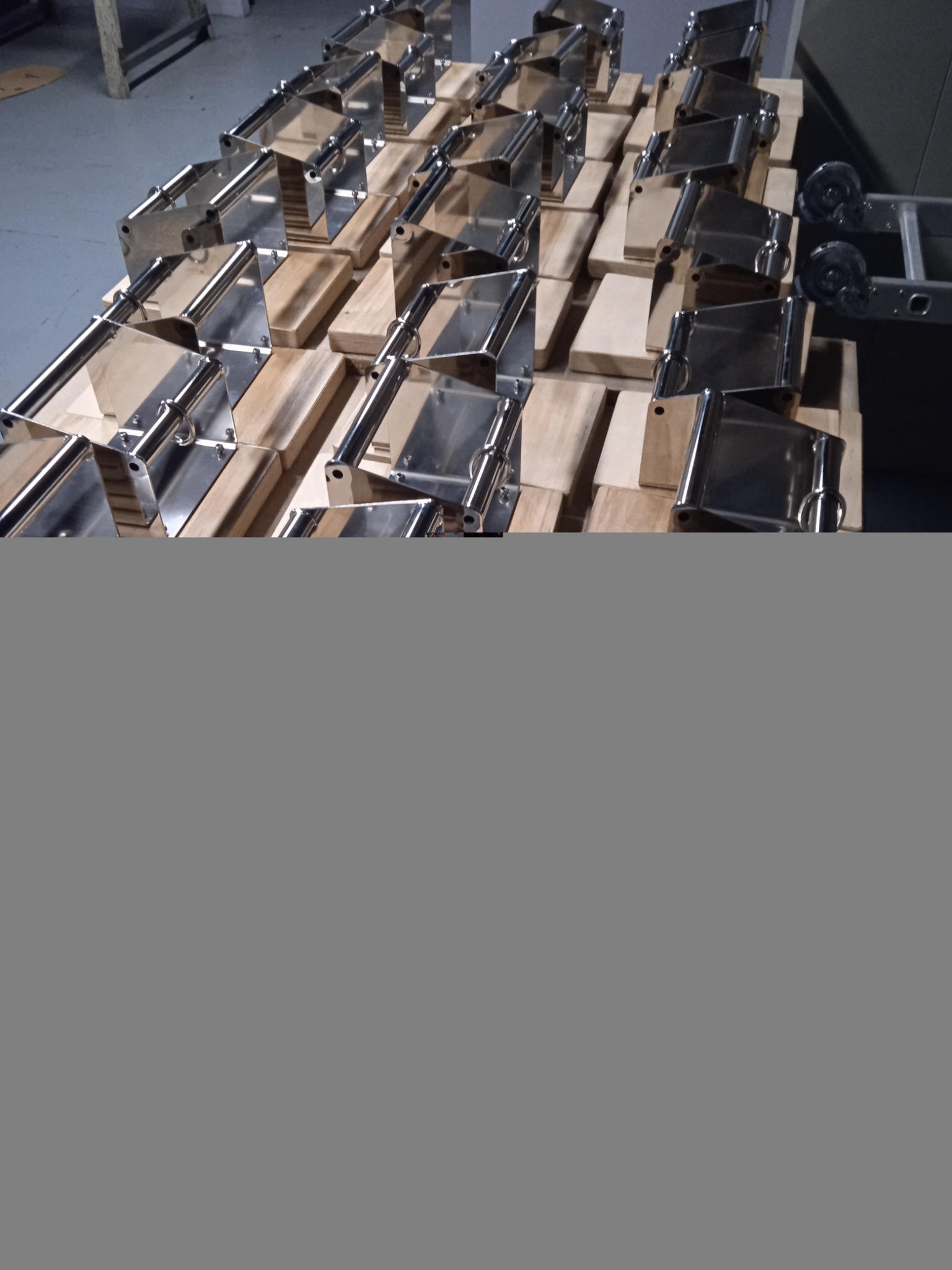 Partia metalowych uchwytów ze stali nierdzewnej, ułożonych na drewnianej palecie w hali produkcyjnej.