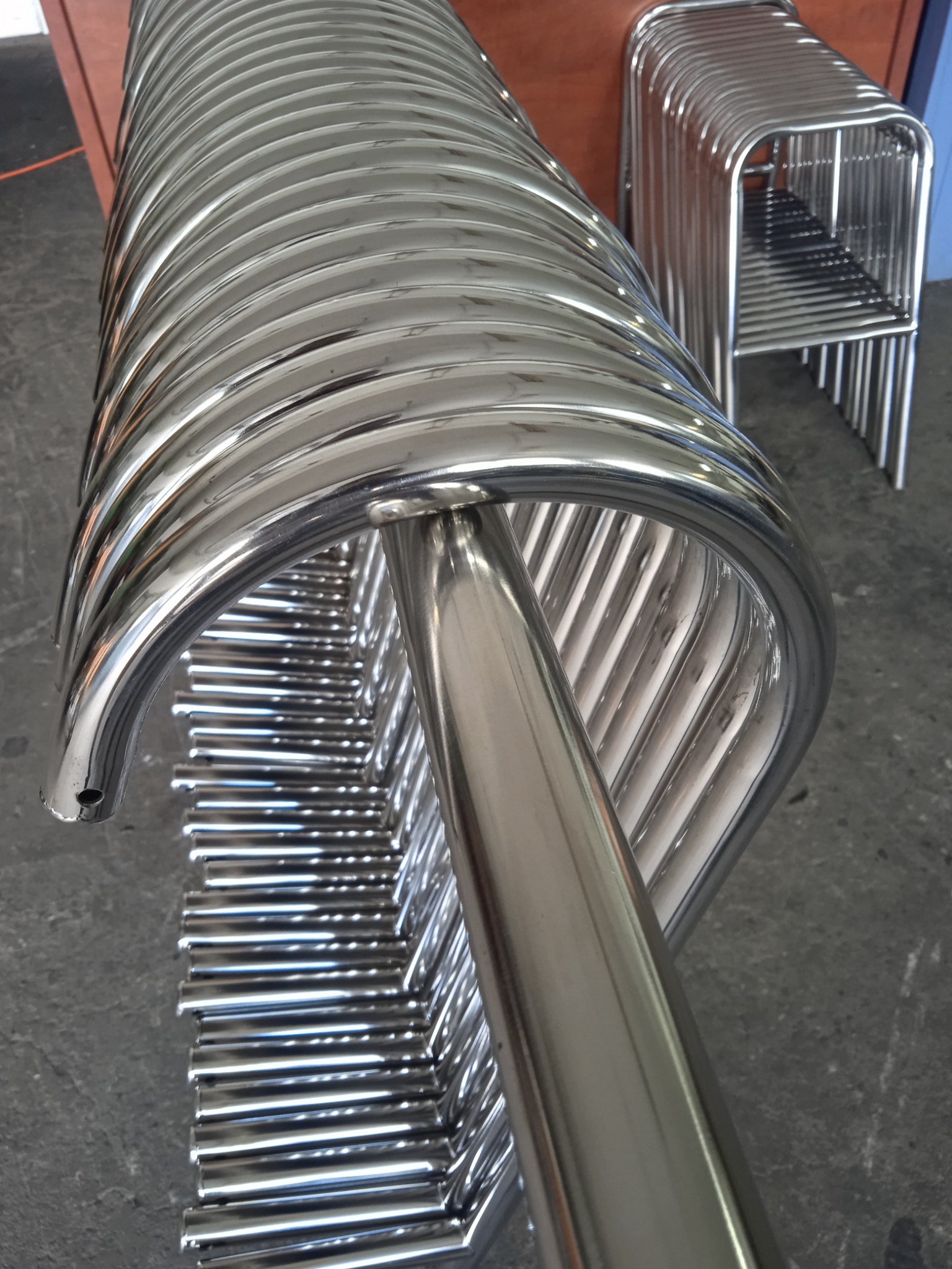 Stos rur giętych ze stali nierdzewnej, ułożonych jedna na drugiej, widoczny otwór na końcu jednej z rur, w tle ułożone w stos metalowe ramy krzeseł