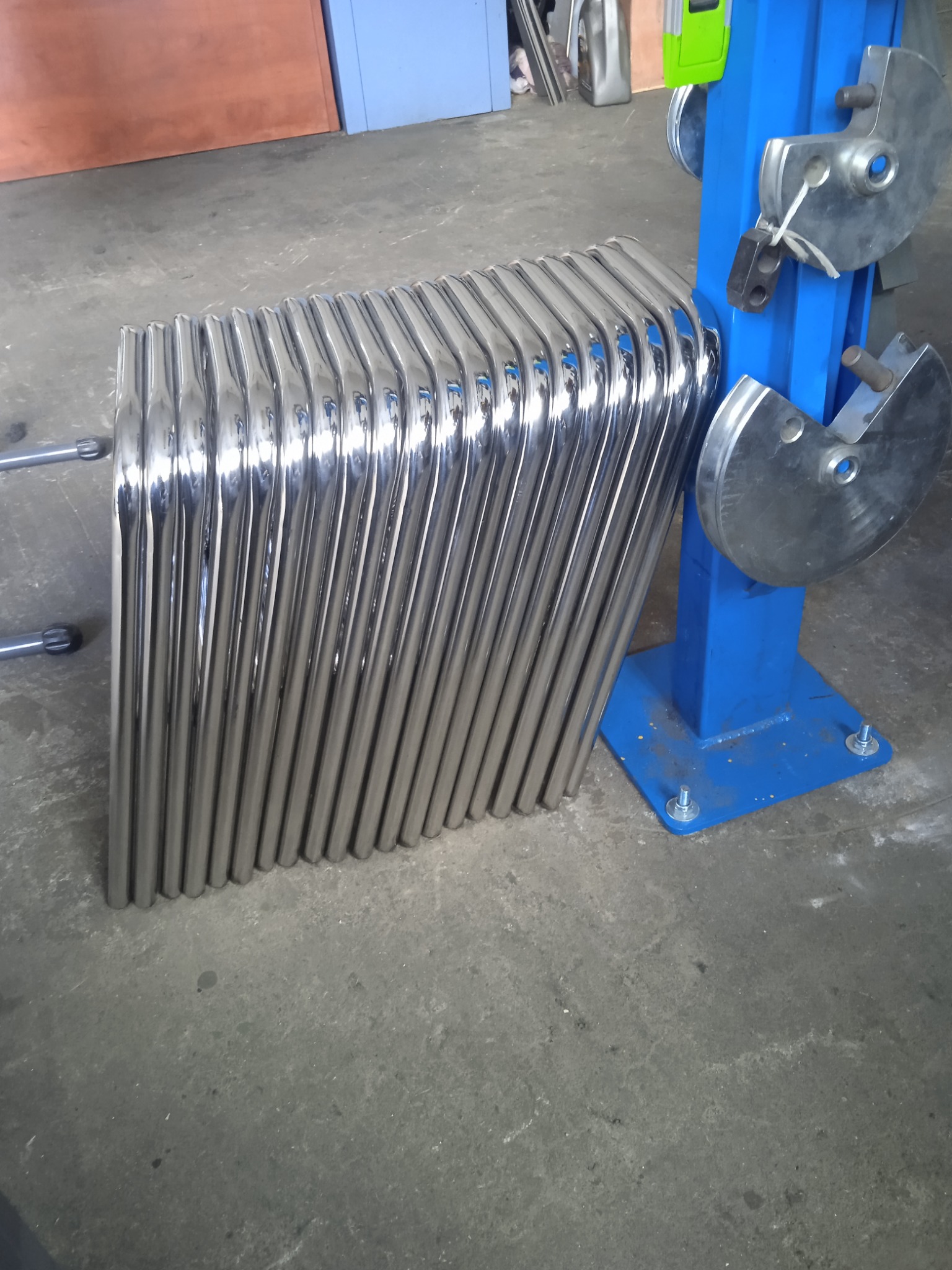 Stos wygiętych, polerowanych rur ze stali nierdzewnej leżący obok niebieskiej giętarki do rur.